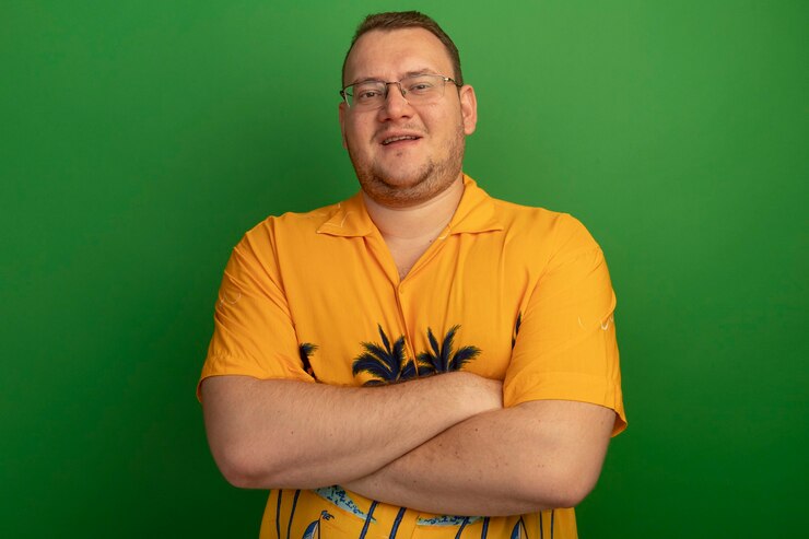 Foto grátis homem de óculos e camisa laranja com sorriso no rosto e braços cruzados em pé sobre a parede verde