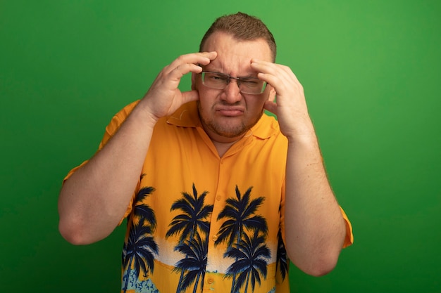Foto grátis homem de óculos e camisa laranja confuso e muito ansioso com as mãos na cabeça em pé sobre a parede verde