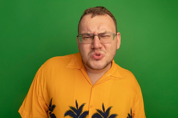 Homem de óculos e camisa laranja descontente com a testa franzida em pé sobre uma parede verde