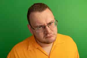 Foto grátis homem de óculos e camisa laranja insatisfeito com uma cara carrancuda em pé sobre uma parede verde
