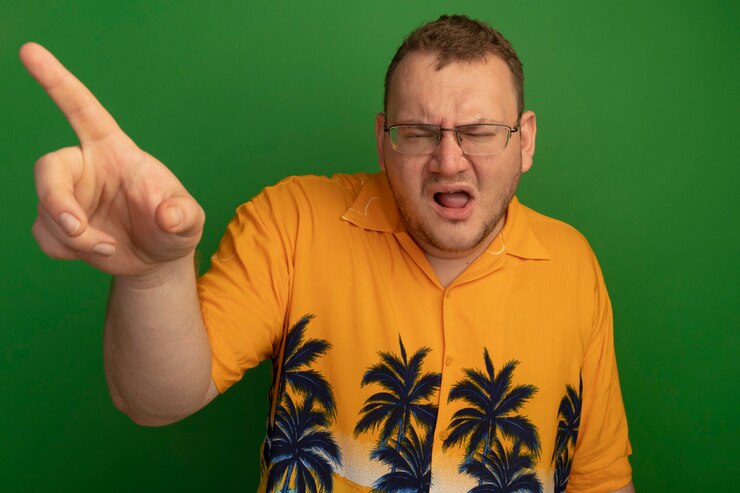 Foto grátis homem de óculos e camisa laranja parecendo confuso e descontente, apontando com o dedo indicador para algo em pé sobre uma parede verde