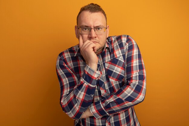 Homem de óculos e camisa xadrez com a mão no queixo pensando com uma cara séria em pé sobre a parede laranja