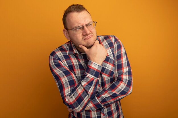 Homem de óculos e camisa xadrez olhando para o lado com uma expressão pensativa no rosto com a mão no queixo pensando em pé sobre a parede laranja