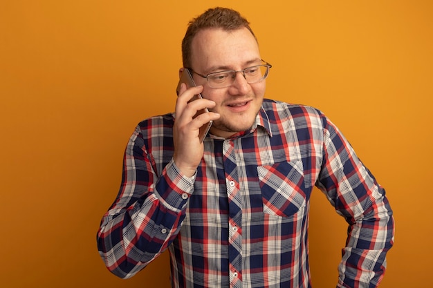 Homem de óculos e camisa xadrez sorrindo enquanto fala no celular em pé sobre uma parede laranja
