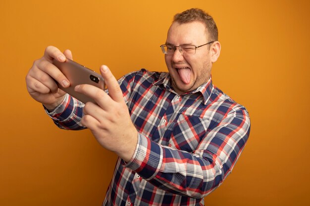 Homem de óculos e camisa xadrez usando smartphine fazendo selfie sorrindo, mostrando a língua em pé sobre a parede laranja