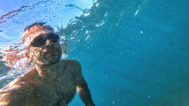Homem de óculos, nadando sob as águas azuis e transparentes do mar Mediterrâneo. Segurando a câmera
