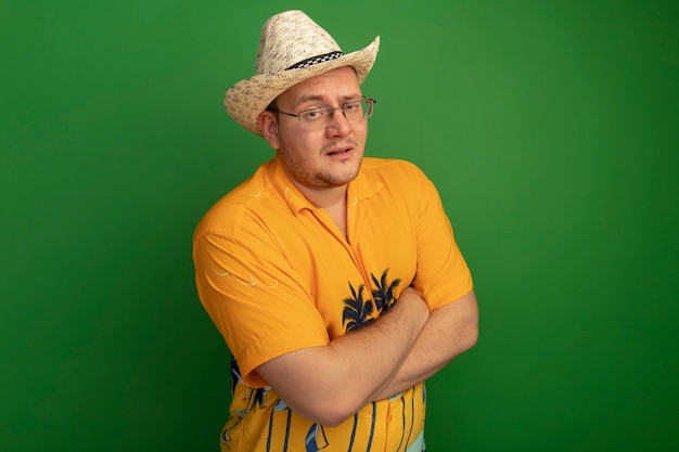 Foto grátis homem de óculos vestindo camisa laranja e chapéu de verão com rosto sério com braços cruzados em pé sobre uma parede verde