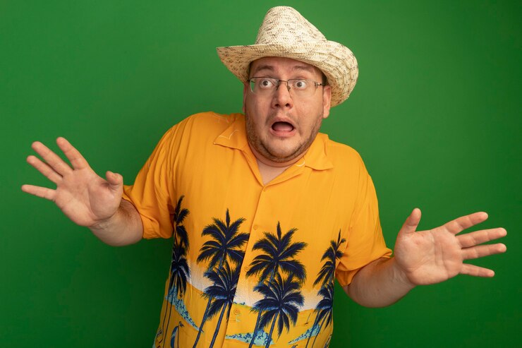 Foto grátis homem de óculos vestindo camisa laranja e chapéu de verão parecendo preocupado e assustado com os braços erguidos em pé sobre a parede verde