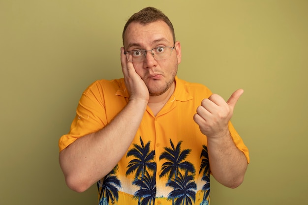 Homem de óculos, vestindo uma camisa laranja, parecendo confuso, mostrando os polegares sendo confundidos em pé sobre a parede de luz