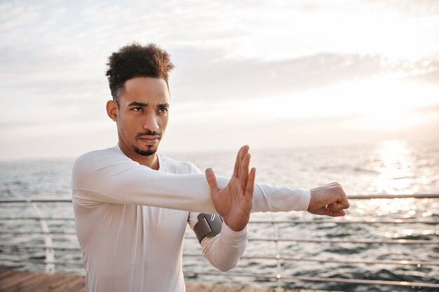 Homem de pele escura, cacheado, autoconfiante e motivado, usando uma camiseta esporte branca de mangas compridas, que se exercita e parece reto perto do mar