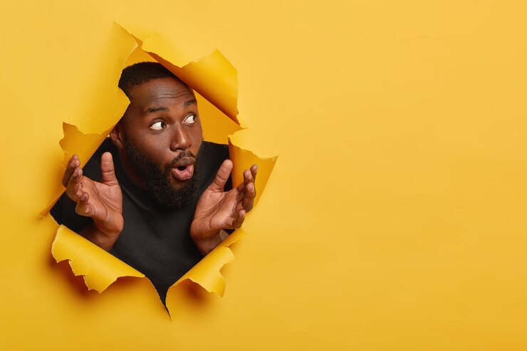 Foto grátis homem de pele escura chocado espalha as palmas das mãos para os lados, reage com surpresa, olha para o lado, veste roupas pretas, focado de lado com expressão impressionada, posa em papel rasgado, fundo amarelo