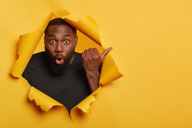 Foto grátis homem de pele escura com a barba por fazer chocado aponta o polegar para longe, sente-se impressionado e atordoado, usa uma camiseta casual preta, fica em um buraco de papel rasgado de fundo amarelo