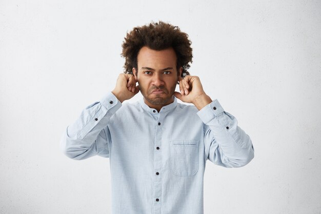 Homem de pele escura com penteado africano curvando os lábios com irritação fechando as orelhas com os dedos