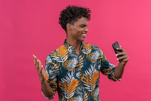 Homem de pele escura, nervoso e zangado, com camisa estampada de folhas, olhando para o celular e levantando a mão