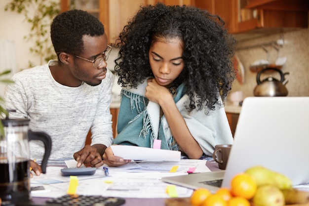 Homem de pele escura segurando um lápis, fazendo cálculos na calculadora e olhando para sua esposa preocupada