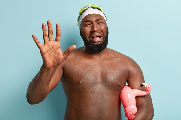 Foto grátis homem de pele escura triste e descontente chora em pânico, mostra a palma da mão, frustrado por ter estragado as férias de verão, usa óculos de proteção, segura ringue de flamingo, isolado na parede azul