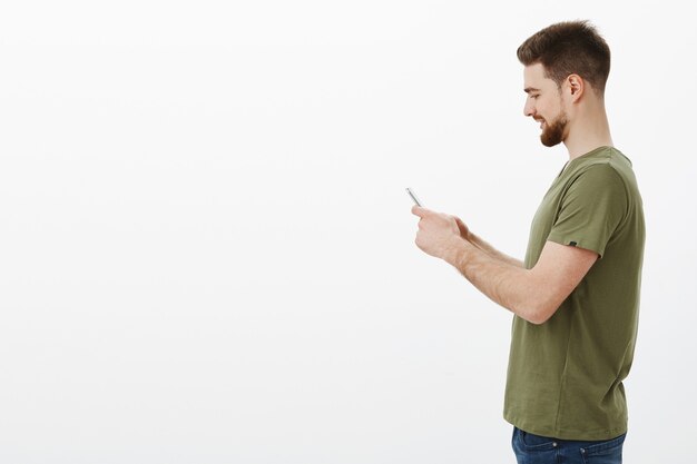 homem de perfil com barba segurando um smartphone e sorrindo animado ao jogar um jogo online interessante via celular, usando a internet para entrar em contato com um amigo, enviando meme legal sobre uma parede branca