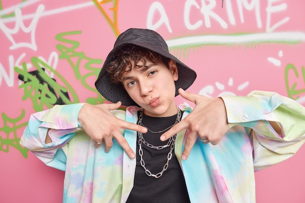 Homem de quatorze anos de idade, um gesto de rap descolado e satisfeito, usando roupas da moda, poses na rua contra uma parede criativa de grafite