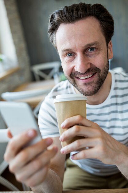 Homem de sorriso que prende o telefone móvel e chávena de café na cafetaria