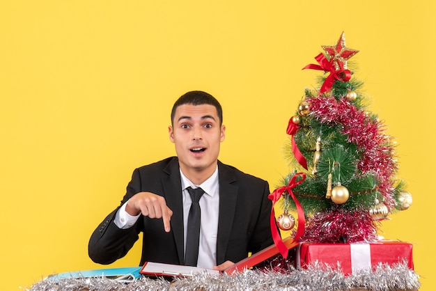 Homem de terno sentado à mesa apontando com o dedo, documento, árvore de natal e presentes