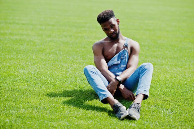 Homem de torso nu afro-americano sexy bonito em macacão jeans sentado na grama verde do campo de futebol do estádio Retrato de homem negro na moda