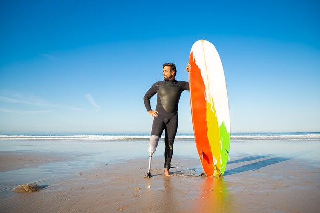 Homem deficiente confiante em pé na praia do mar com prancha