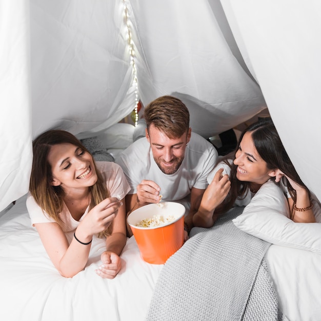 Homem deitado com duas amigas na cama comendo pipocas