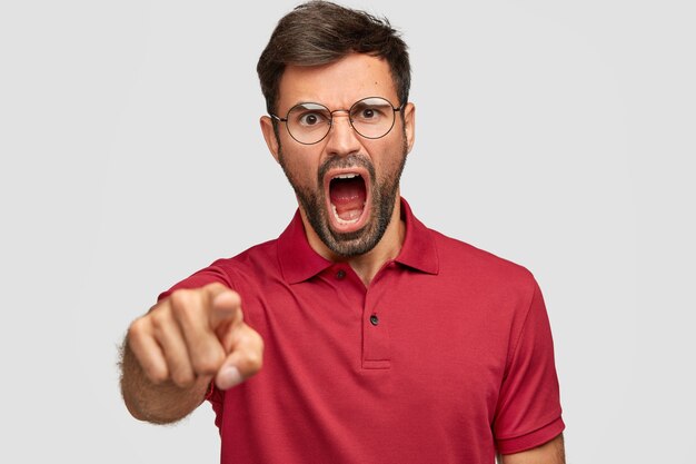 Homem deprimido furioso com cerdas escuras, grita com raiva de alguém, pontos, vestido com camiseta vermelha brilhante, isolado sobre a parede branca. Homem louco com a barba por fazer expressa raiva, grita bem alto