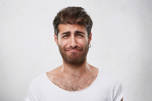 Homem desanimado com penteado e barba franzindo a testa, lamentando o que fez. homem entristecido em uma camiseta branca. pessoas, moda, estilo de vida, conceito de emoções