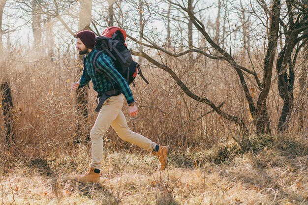 Homem descolado e moderno viajando com uma mochila na floresta de outono, usando chapéu e camisa quadriculada, turista ativo correndo, explorando a natureza na estação fria