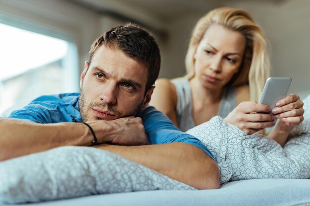 Homem descontente deitado na cama e ignorando sua namorada que está enviando mensagens de texto no celular