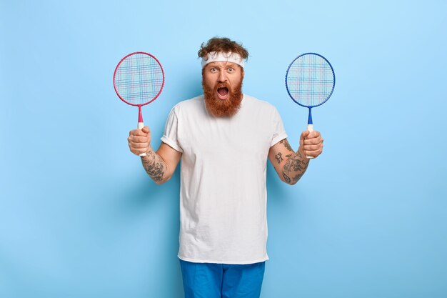 Homem desportivo indignado segura duas raquetes de badminton, o amigo zangado não apareceu no jogo
