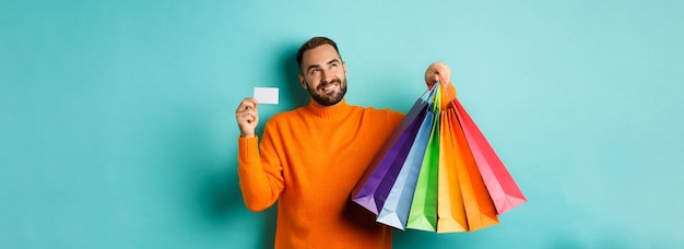 Homem devidamente feliz mostrando cartão de crédito e sacolas de compras em pé contra fundo turquesa