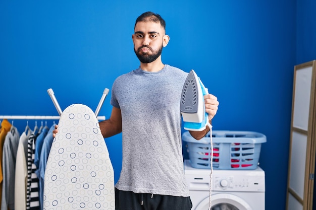 Foto grátis homem do oriente médio com barba segurando bochechas de ferro elétrico com cara engraçada boca inflada com ar pegando ar