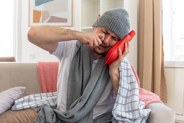 homem doente a chorar com um lenço no pescoço e chapéu de inverno, segurando e colocando a cabeça em uma garrafa de água quente, sentado no sofá da sala de estar