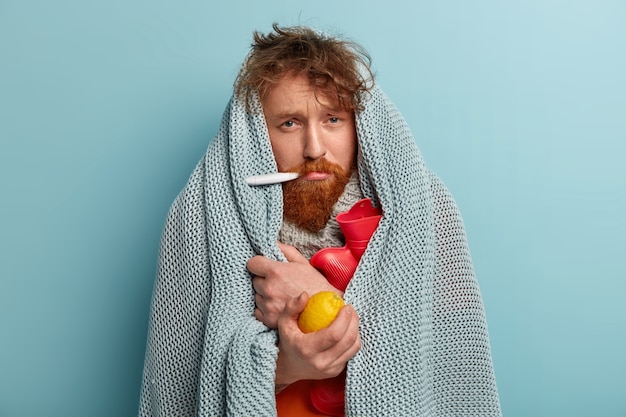 Homem doente com roupas quentes com termômetro, segurando limão, bolsa de água quente