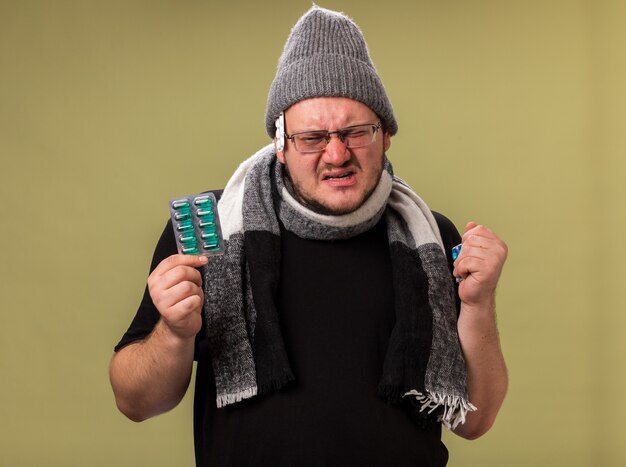 Homem doente de meia-idade olhando para a câmera com chapéu de inverno e lenço segurando comprimidos