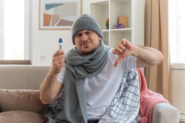 homem doente e desagradável com um lenço no pescoço, um chapéu de inverno segurando um termômetro e sentado no sofá da sala com o polegar