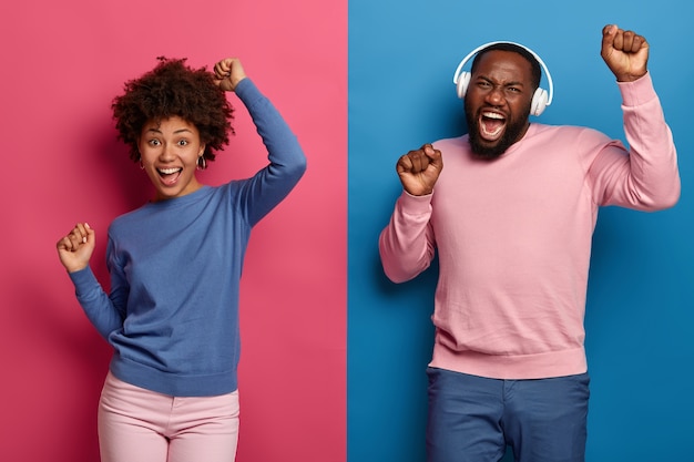 Homem e mulher afro-americana alegre carismática levantam as mãos e dançam alegremente com o ritmo da música, usam fones de ouvido, posam contra o espaço azul e rosa. Pessoas