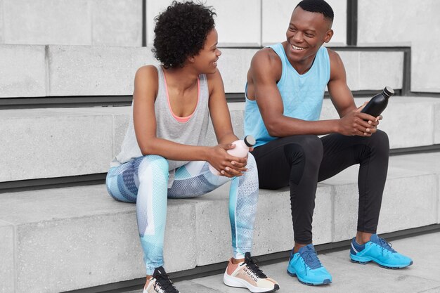 Homem e mulher de pele escura, alegre e motivada sentam-se na escada, olham positivamente um para o outro, seguram garrafas com água, usam roupas esportivas e tênis, discutem sobre competições esportivas.