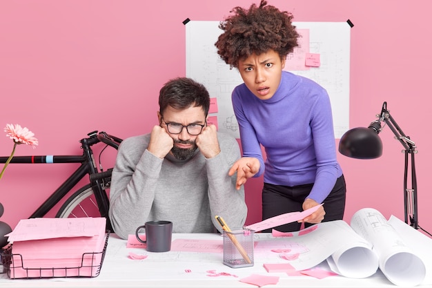 Homem e mulher habilidosa e descontente e intrigada colaboram em esboços de design em pose de espaço de coworking para criar plantas vestidas com roupas casuais. Colegas de raça mista preparam apresentação ou projeto