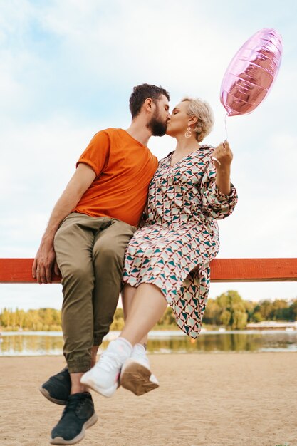 Homem e mulher se beijando sentados e segurando um balão de coração rosa com o céu