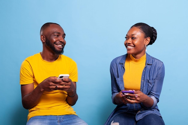 Foto grátis homem e mulher segurando smartphones e sorrindo um para o outro, usando telefones celulares com tela sensível ao toque para navegar na rede de internet. casal moderno rindo e usando conexão de mídia social em estúdio.