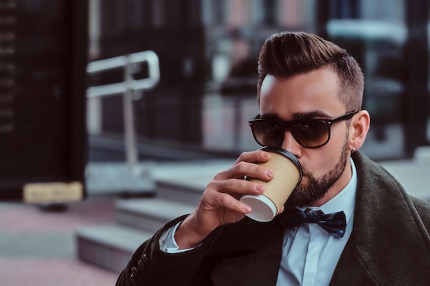 Homem elegante atraente em óculos de sol está bebendo seu café enquanto está sentado lá fora na cafeteria.