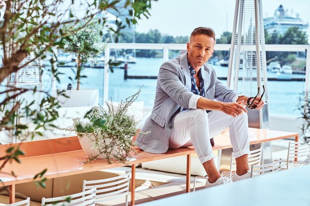 Homem elegante bem sucedido vestido com roupas elegantes modernas, sentado em uma mesa no café ao ar livre no contexto do cais da cidade.