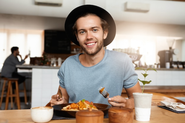 Homem elegante com barba para aplacar a fome enquanto janta sozinho em um restaurante moderno em um dia ensolarado, comendo uma refeição com garfo e faca