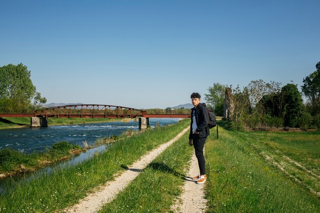 Homem elegante jovem de pé no caminho perto do rio com mochila de transporte