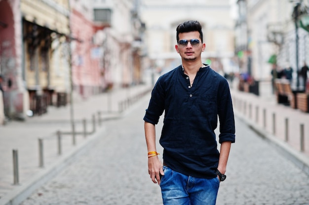 Homem elegante modelo indiano em roupas casuais, camisa preta e óculos de sol posados ao ar livre na rua da Índia