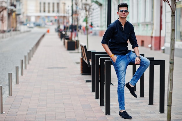 Homem elegante modelo indiano em roupas casuais, camisa preta e óculos de sol posados ao ar livre na rua da Índia