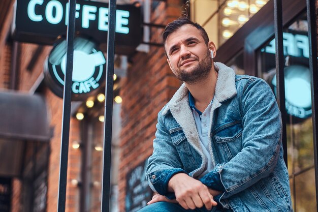 Homem elegante sorridente, vestindo uma jaqueta jeans com fones de ouvido sem fio, segurando o café para viagem fora do café.